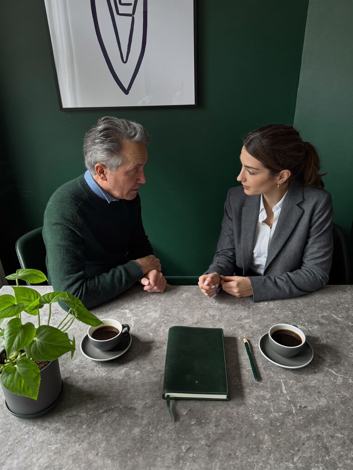 Zwei Personen im Gespräch an einem Steintisch vor dunkelgrüner Wand, dazwischen ein Notizbuch und Kaffeetassen.